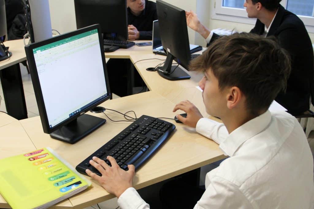 Jeune qui travaille sur un tableau Excel devant son écran d'ordinateur en salle informatique.
